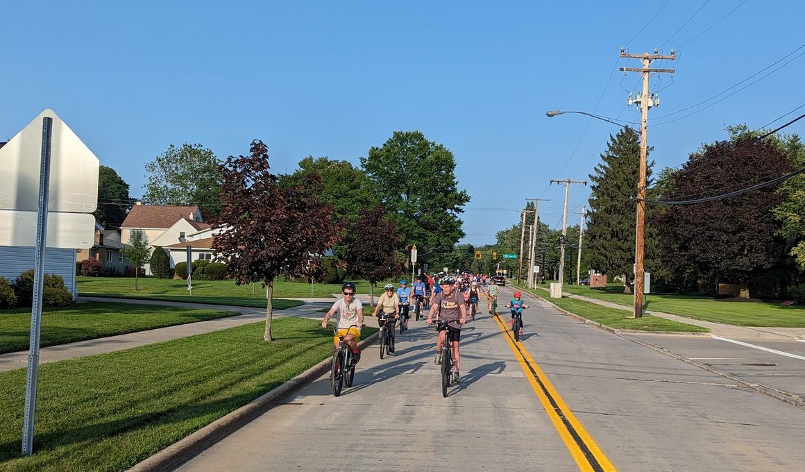 A line of Bike Solon riders of all ages, led by Solon's mayor in a Bike Solon shirt, heading down a Solon residential street, following the road's double-yellow centerline