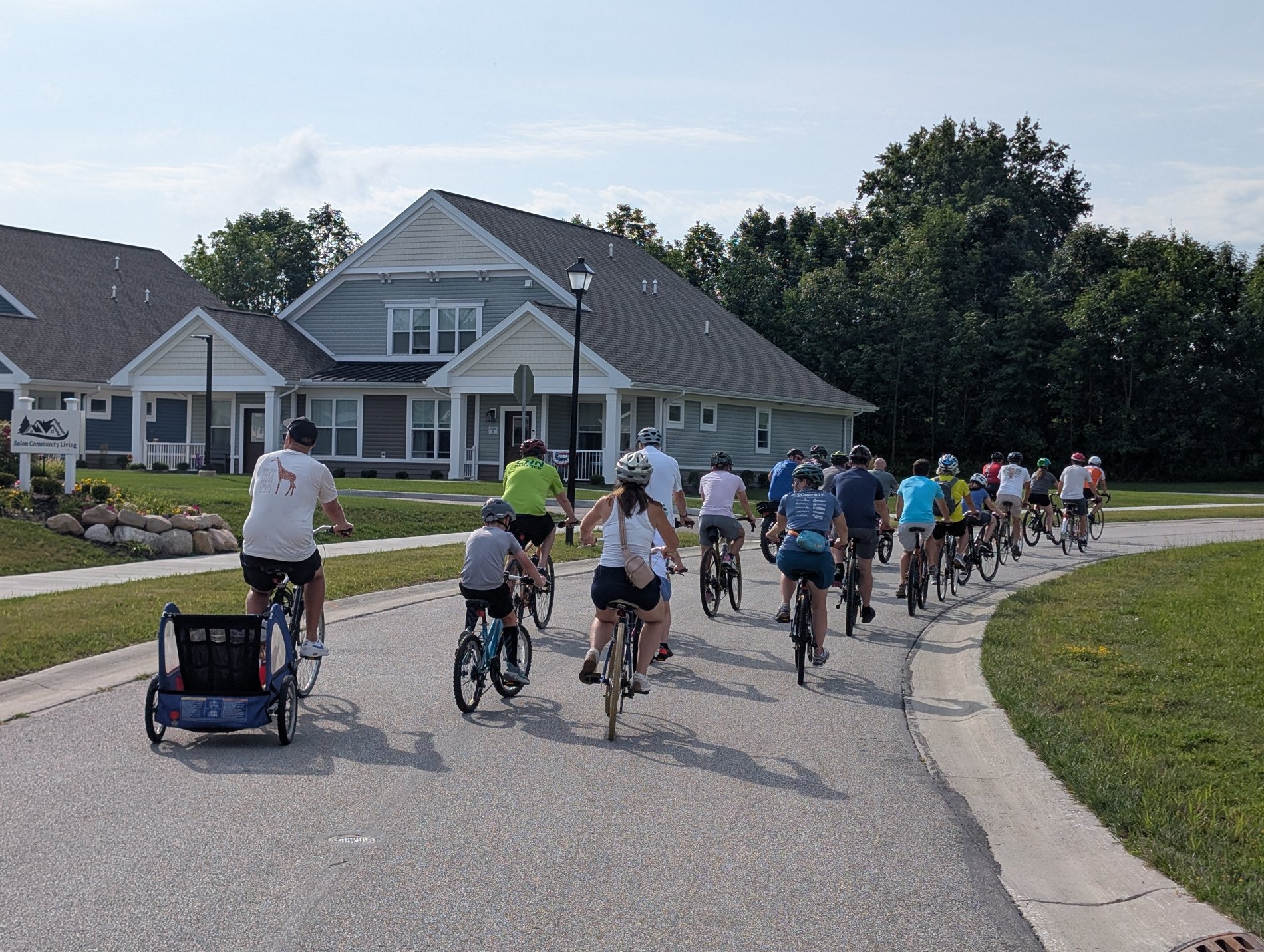 A family with a bike trailer leads a line of riders down a neighborhood street past Solon Community Living